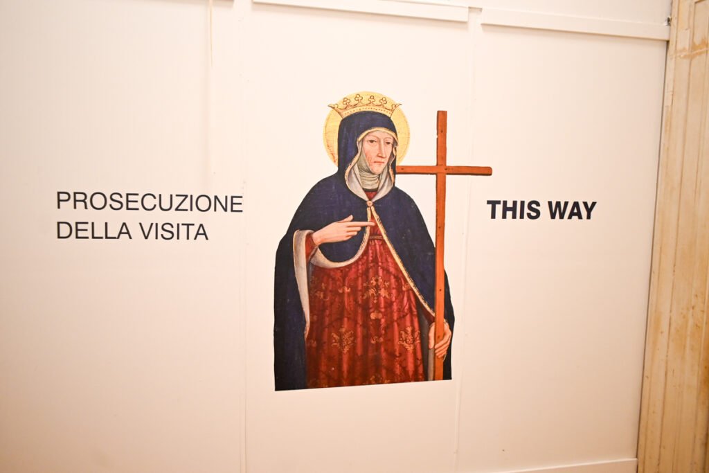 Hintergrund: Ein Wandbild mit der Darstellung einer Heiligenfigur, die an die christliche Kunst erinnert, und Schilder mit Anweisungen auf Italienisch und Englisch.